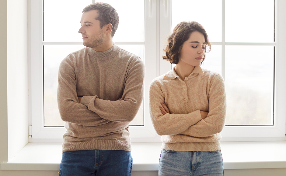 A man and woman with folded arms sit side by side on a windowsill, looking away from each other with neutral expressions.