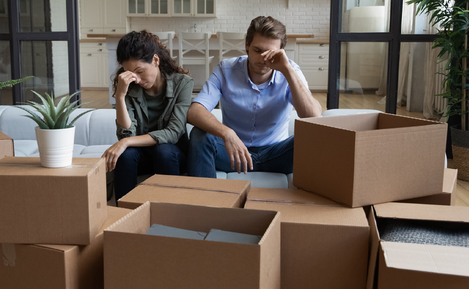 Two people sit on a couch looking stressed, surrounded by unpacked cardboard boxes in a home setting.