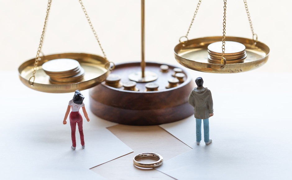 Miniature figures of a man and woman stand on opposite sides of a balance scale with coins, symbolizing divorce and asset division. A wedding ring lies in front.