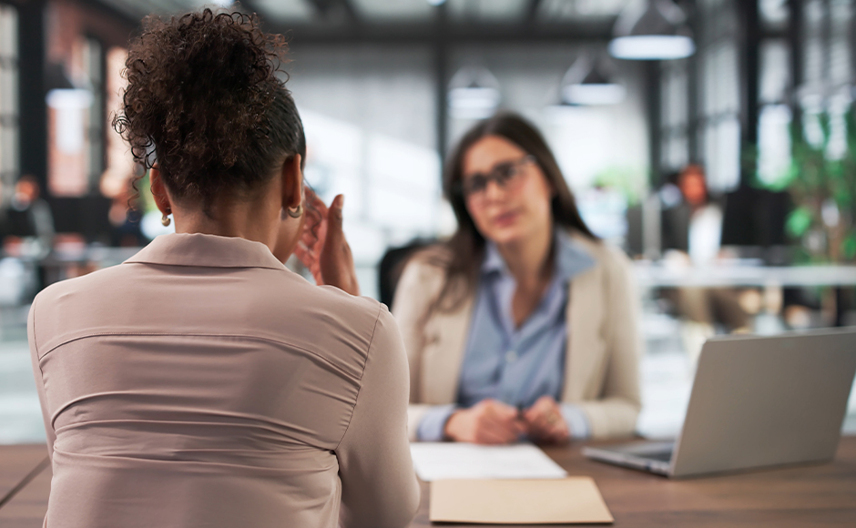 Two women meet in a bright office: one gestures with her back to the camera while the other listens beside an open laptop, creating a professional tone.