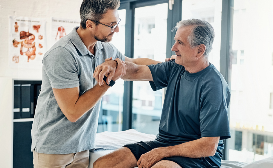 A healthcare professional assists an older man with arm exercises during a physical therapy session in a medical office.