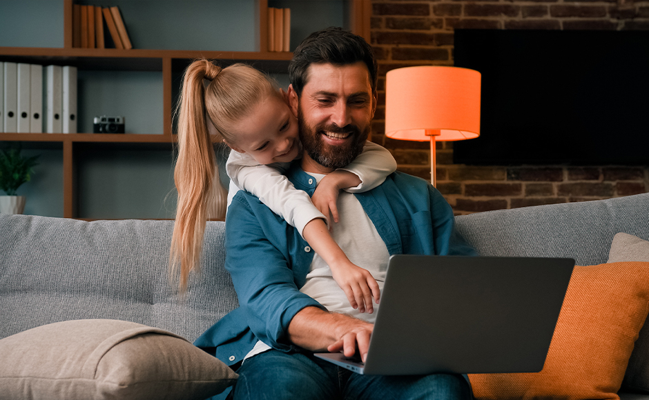 Father working on a laptop smiles warmly as his daughter hugs him from behind on a cozy couch. A soft lamp glows in the homey background.