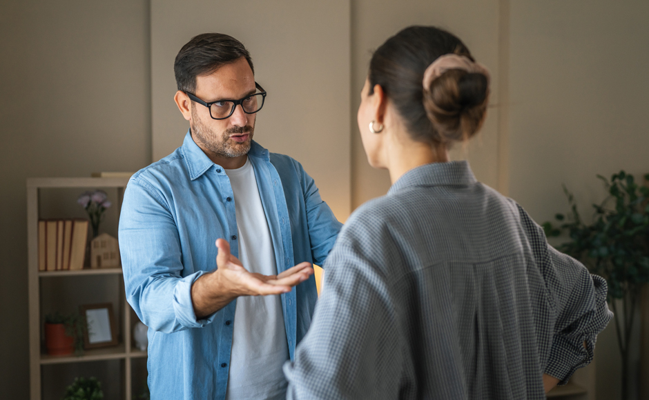 A man in glasses and a woman converse in a cozy room with shelves, plants, and warm lighting. The man gestures expressively, appearing thoughtful.