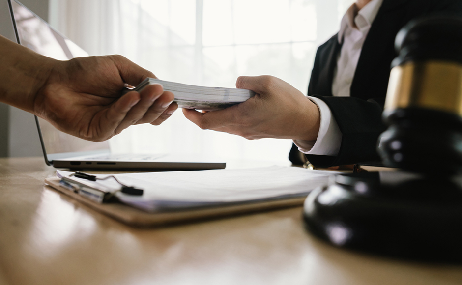 Two people exchange a stack of cash across a table with a clipboard and laptop. A judge's gavel is visible, suggesting a legal or business context.