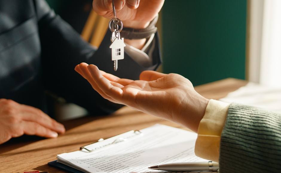 Close-up of a person handing house keys with a house-shaped keychain to another person over a signed contract on a wooden desk, conveying a sense of accomplishment.