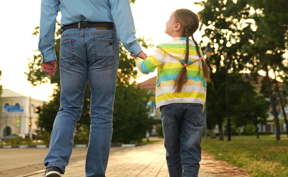 An adult and a child holding hands walk on a paved path in a park. The child has braided hair and is looking up at the adult. Trees and buildings are visible in the background.