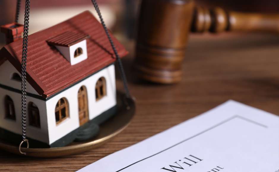 A model house sits on a scale next to a gavel and a document titled 'Will' on a wooden table, conveying themes of inheritance and legal decisions.