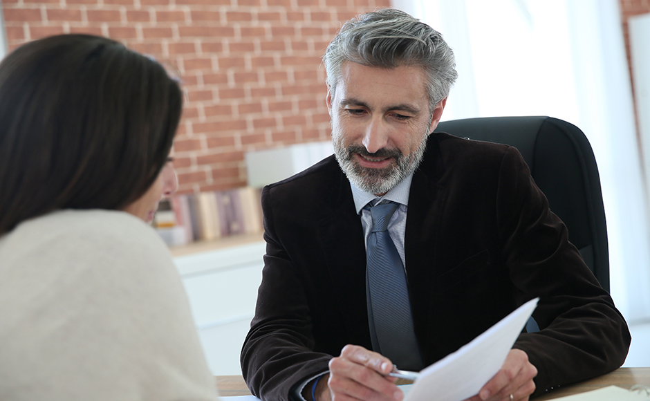 A bearded man in a suit sits at a desk, holding papers and smiling at a woman across from him. The setting is a modern office with brick walls.