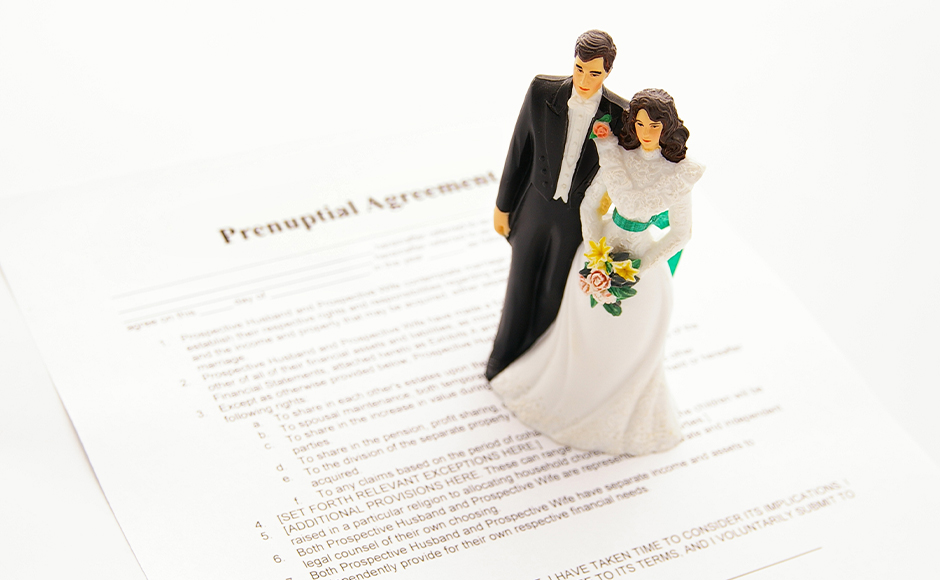 A bride and groom cake topper stands on a prenuptial agreement. The couple is dressed formally; the setting suggests themes of marriage and legal preparation.