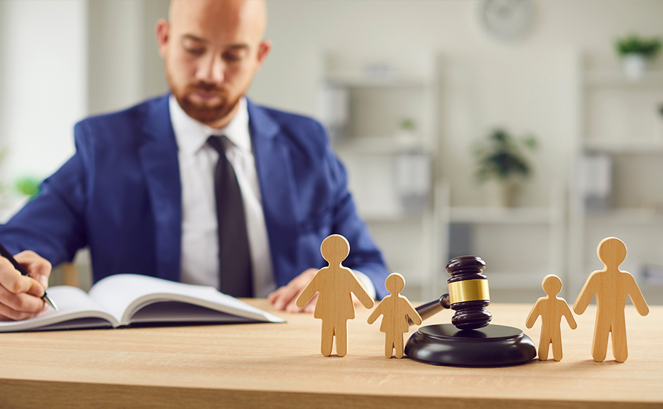 A man in a suit writes in a book at a desk with wooden family figures and a gavel nearby, symbolizing legal family matters. The mood is serious and professional.